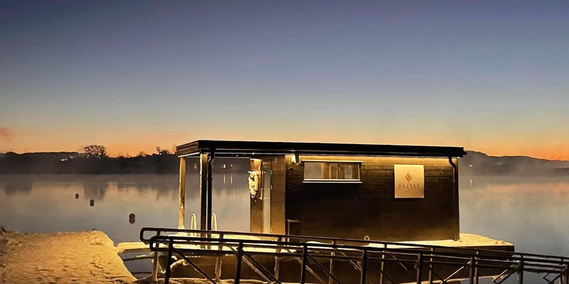 Flytende sauna ved vannet en vinterkveld, omgitt av snø og stille vannspeil.